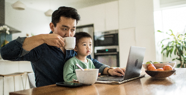 A father takes a sip of coffee while seated at the table with his young son in his lap and his laptop open in front of him.